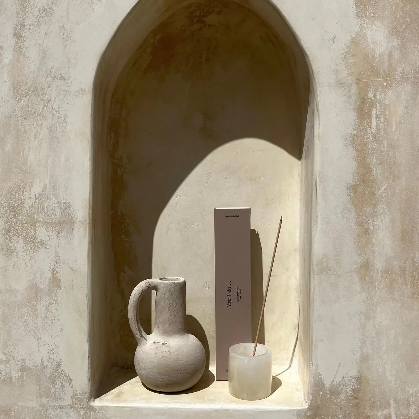 A rustic alcove with a textured beige wall displays a ceramic jug, a glass with burning incense casting soft shadows, and Nusa Holistick's Essential Oil Incense—Jasmine, Lotus, Champaka—in its tall handcrafted box.
