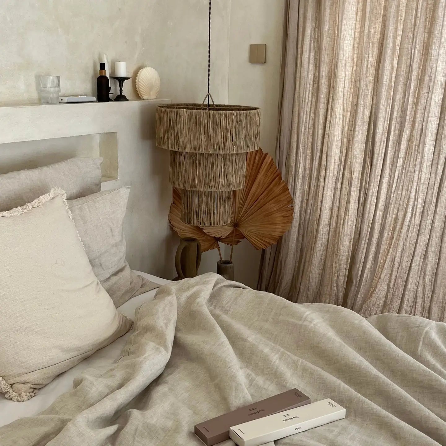 A cozy, neutral-toned bedroom with a rumpled bed, beige linens and pillows. Nusa Holistick Essential Oil Incense (Jasmine, Lotus, Champaka) fills the air. Soft light filters through sheer beige curtains; two long boxes rest on the bed.