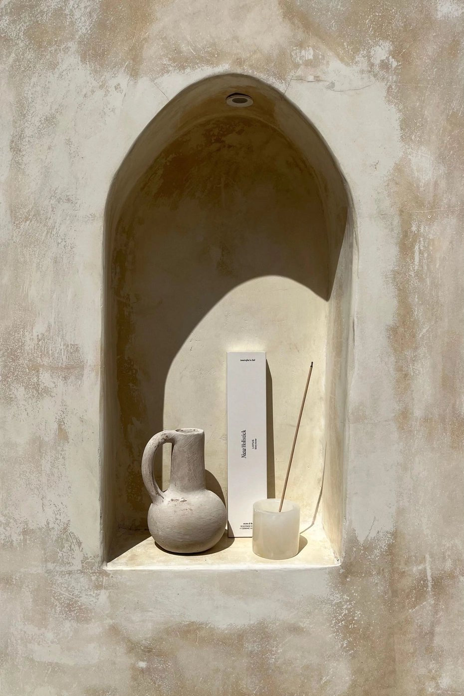 A recessed wall niche displays a rustic ceramic pitcher, a white box, and a glass container of Nusa Holistick Essential Oil Incense (Jasmine, Lotus, Champaka), all illuminated by natural light against a textured beige wall.
