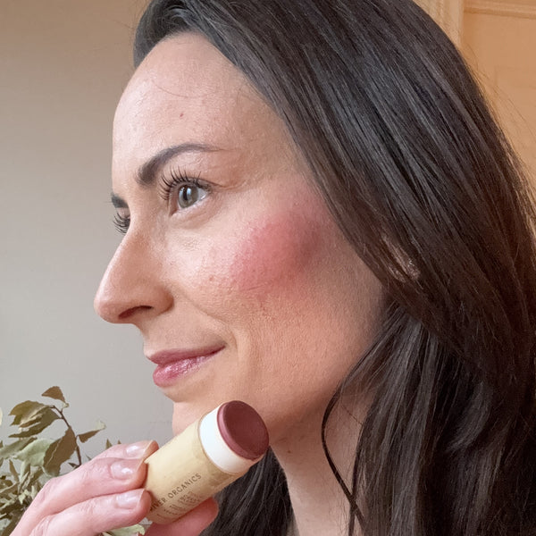 A person with long dark hair holds the River Organics Creamy Vegan Blush Stick - Pinch against their cheek. The wooden tube accentuates its vegan, cruelty-free formula. A softly lit plant-filled background emphasizes the product's natural essence.