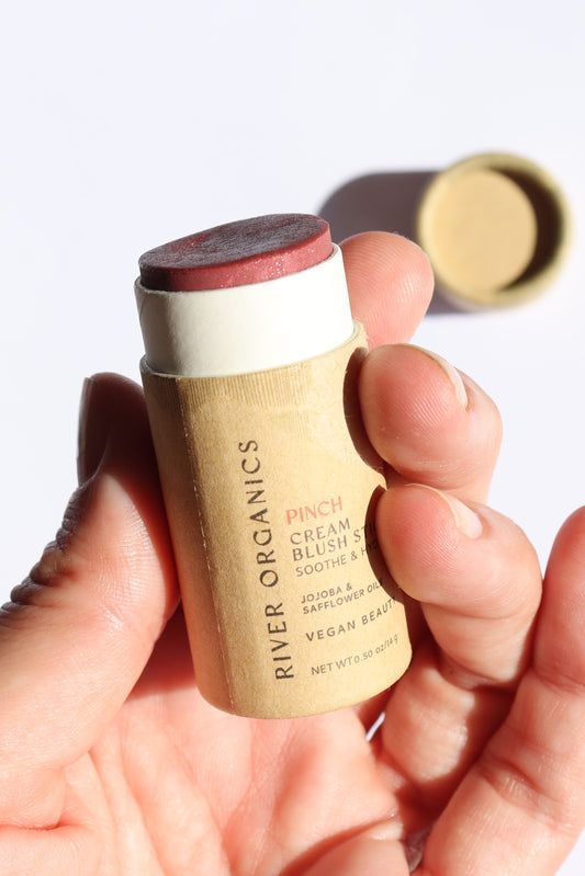A hand holds an open River Organics Natural Cream Blush Stick | Vegan + Multi-Use in a rosy red shade. This zero-waste blush comes in eco-friendly, cylindrical packaging—an ideal plastic-free, vegan, and organic makeup choice.