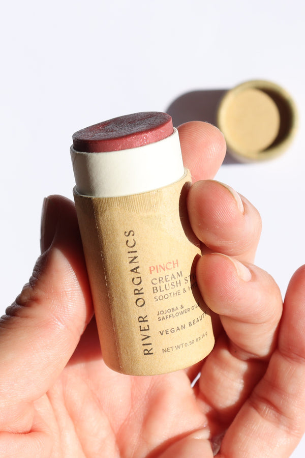 A hand holds the River Organics Natural Cream Blush Stick | All Colors, featuring a reddish-pink tint in a zero-waste cardboard tube. The plastic-free cap rests nearby on a white surface.