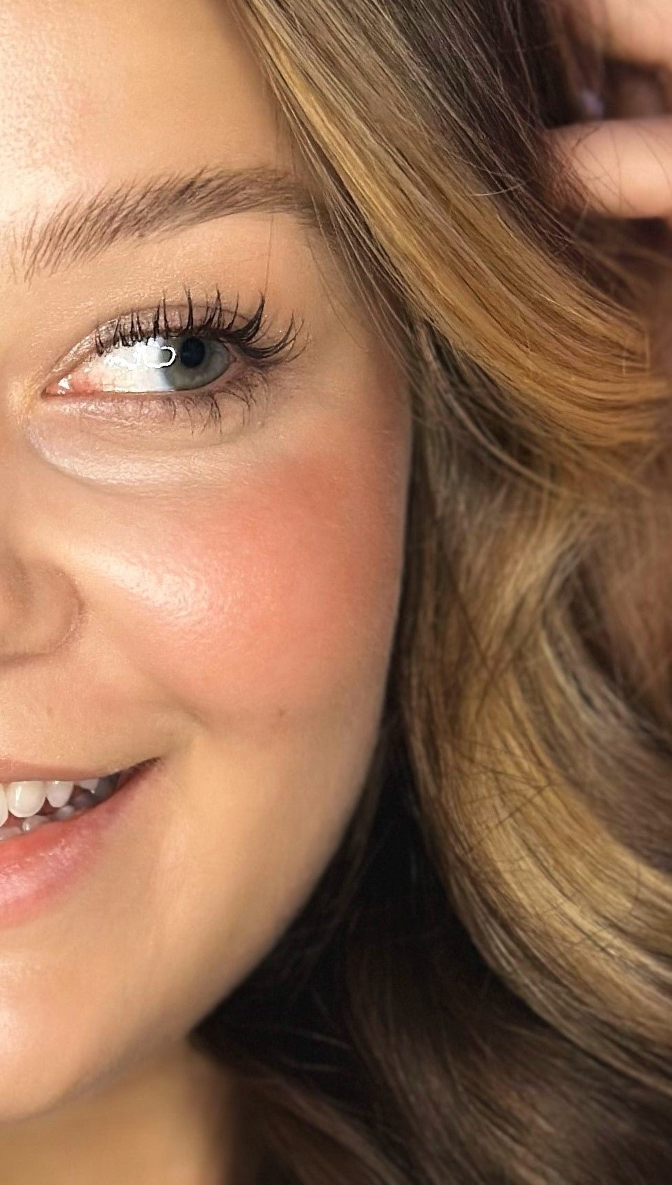 A close-up of a woman’s serene face highlights her eye, cheek, and smile. Her skin looks smooth with River Organics' Creamy Vegan Blush Stick in Pinch adding a natural glow to her cheeks. Light brown hair frames a face contentedly enhanced by nourishing ingredients.