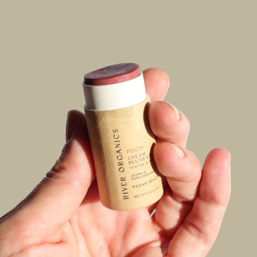 A hand holds a River Organics Creamy Vegan Blush Stick - Pinch, showcasing a red tint and eco-friendly packaging. The label highlights its soothing, vegan, cruelty-free, zero-waste formula with skin-nourishing ingredients, set against a neutral beige background.