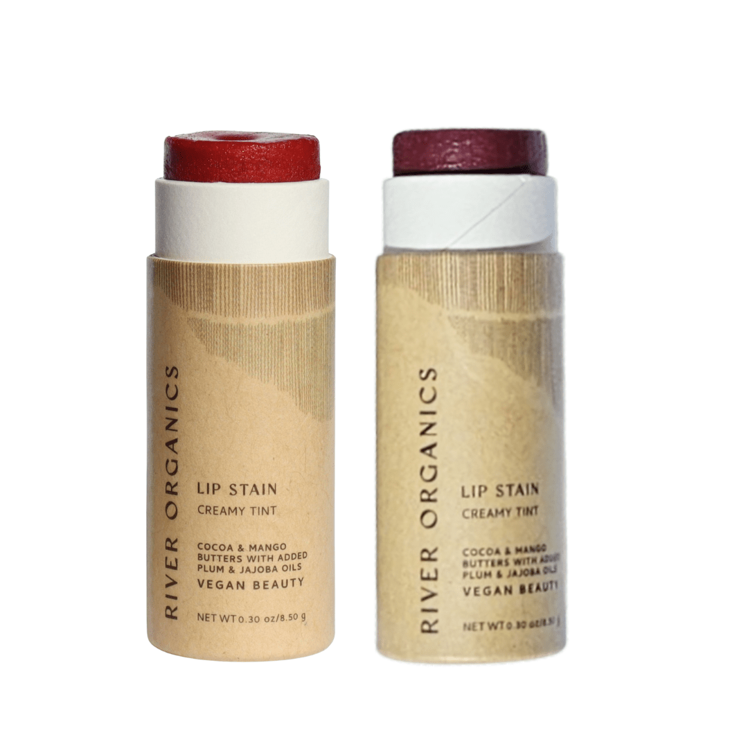 Two tubes of River Organics Lip Stain Sets in cardboard packaging, tops open to show red and plum non-toxic tints. Labels highlight vegan, natural formulas from River Organics.