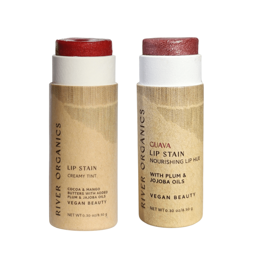 Two River Organics Lip Stain Sets in cardboard tubes stand upright, labeled "Creamy Tint" and "Guava Lip Stain." Both feature non-toxic, vegan formulas with nourishing natural oils.