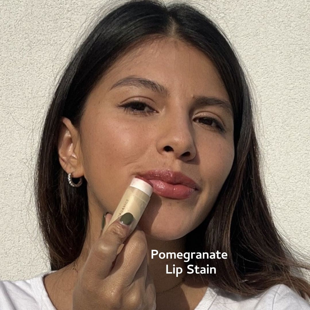 A woman with long dark hair applies River Organics Vegan Lip Stains from a tube to her lips, standing against a light wall. The text "Pomegranate Lip Stain" appears on the image.