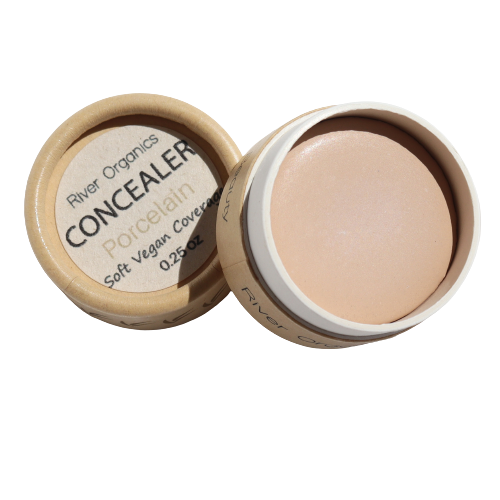 An open container of River Organics' Imperfect Natural Hydrating Vegan Concealer in Porcelain shows the creamy formula. The zero-waste tube, labeled "Soft Vegan Coverage" and "0.25 oz," features nourishing organic plant oils.