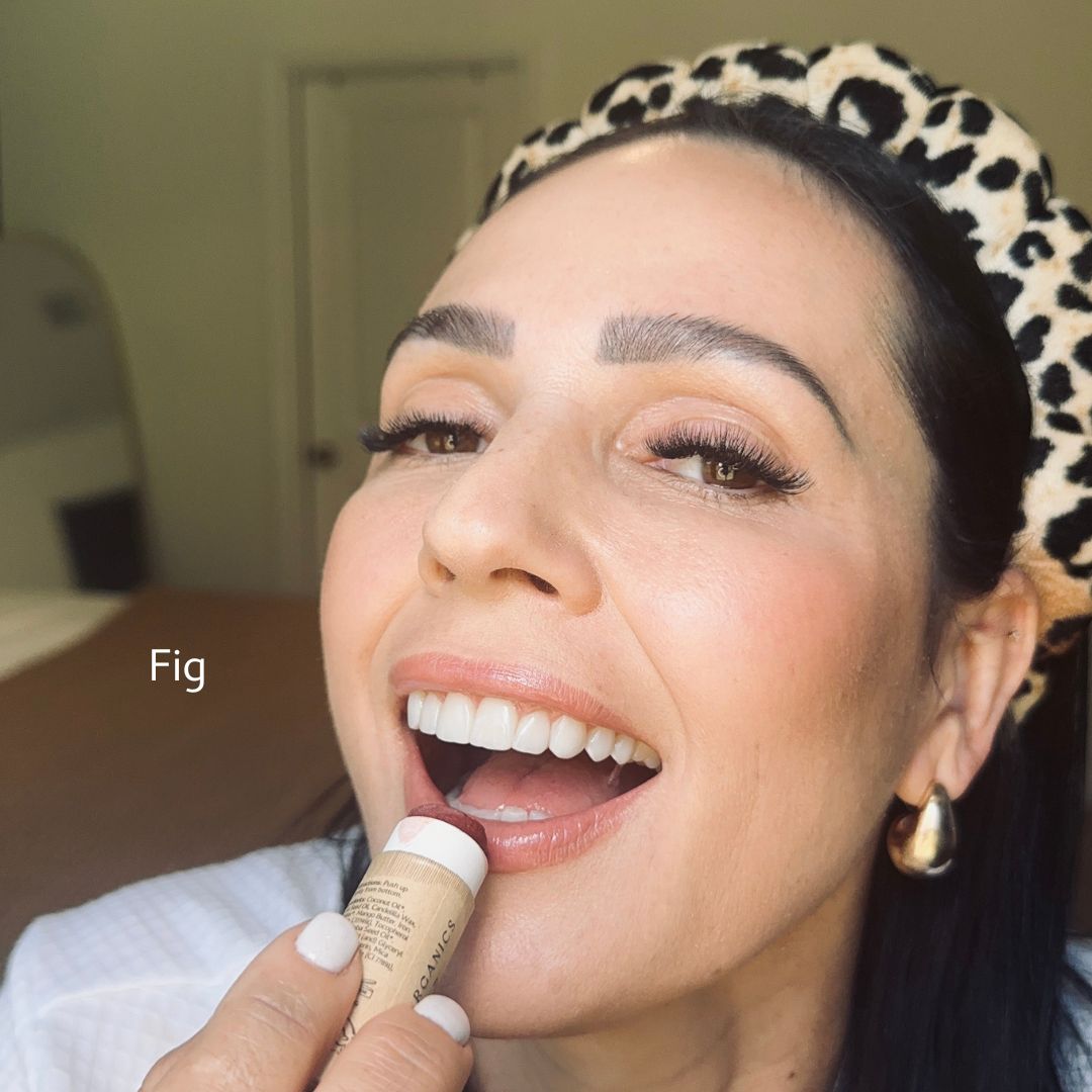 A smiling woman with dark hair and a leopard-print headband applies River Organics Vegan Lip Stains in "Fig." She has flawless makeup, wears gold earrings, and the word "Fig" appears beside her face.