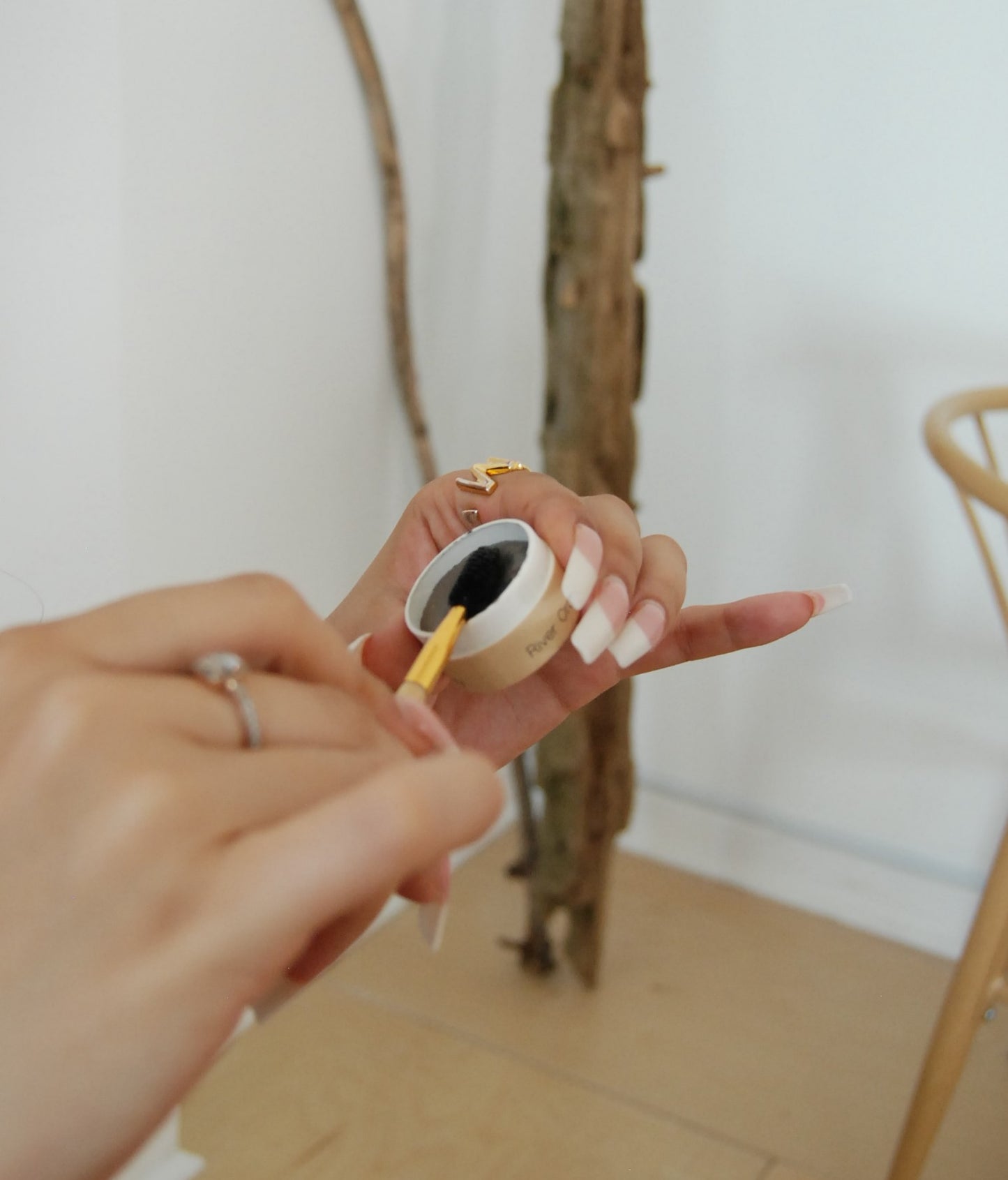 A person with a ring and long manicured nails is using a small brush to apply black powder from a small, round container of River Organics Tinted Vegan Eyebrow Wax | Dark. The background features a light-colored wooden floor and a piece of leaning driftwood.