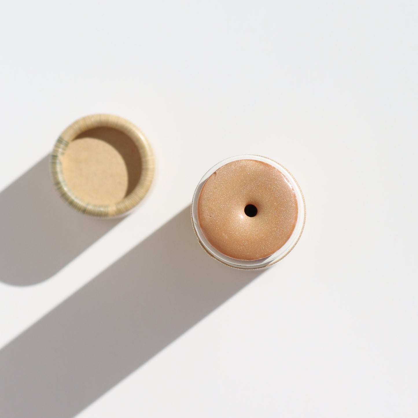 A top view shows the Illumi Vegan Highlighter Stick - Golden Hue by River Organics, revealing a beige, creamy substance inside its open cylindrical container. It casts a shadow on the white surface beside its lid, highlighting its sustainable packaging design.