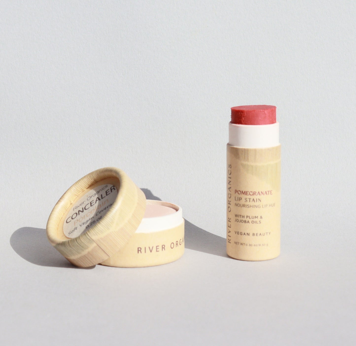 A small open pot of River Organics’ zero-waste vegan concealer and a tube of their pomegranate lip stain, both from the Lip + Concealer Set and presented in light, eco-friendly packaging, are placed on a neutral surface with soft lighting—perfect additions for any clean beauty routine.