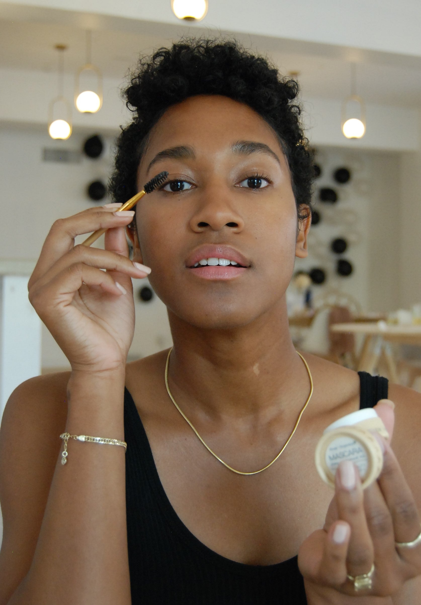 A person with short curly hair is applying River Organics Black Zero-Waste Mascara to their eyelashes using a small brush. They are holding a container of makeup and wearing a black tank top, a necklace, and a bracelet. The background appears to be a brightly lit, minimalist room with white walls.