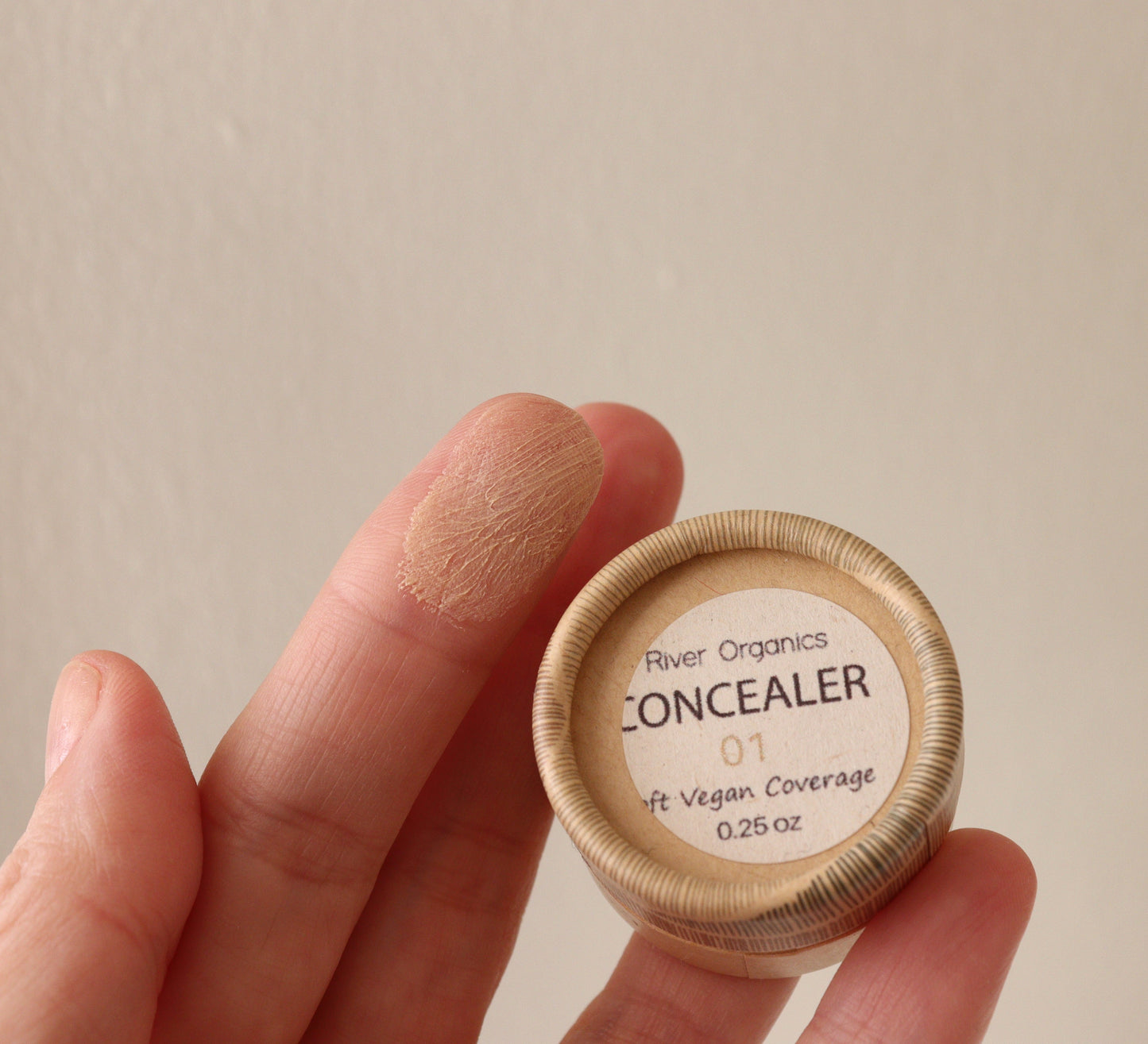 A hand holds a small container of River Organics Titanium Dioxide-Free, Zero-Waste Vegan Concealer with shade 01 swatched on the index finger. The label reads "Vegan Coverage" and "0.25 oz." against a plain, neutral background.
