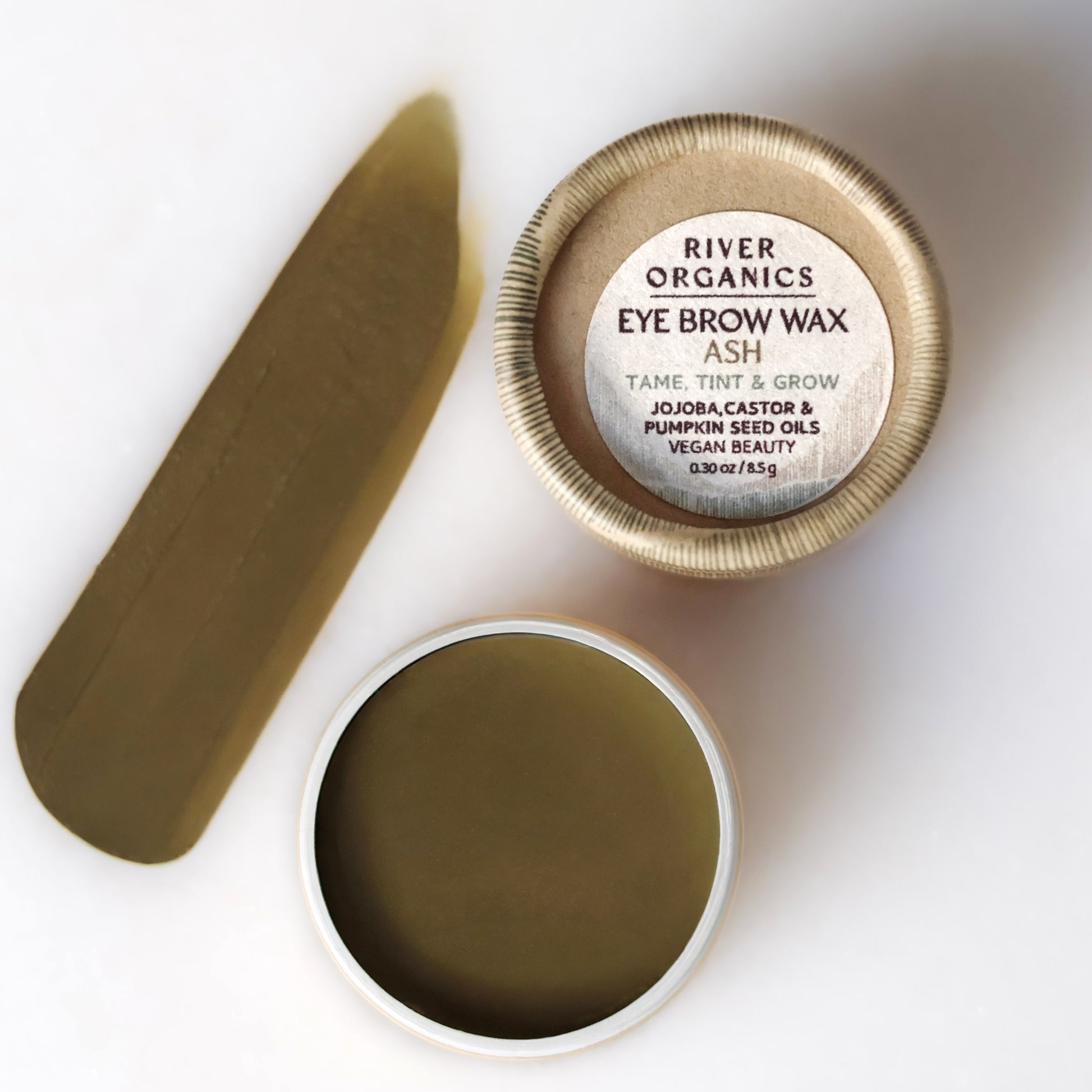 A round container of River Organics Brow Wax & Growth Serum sits open, displaying olive-green wax. Next to it is a swatch and the lid featuring product details. This vegan brow product offers both styling and growth benefits.
