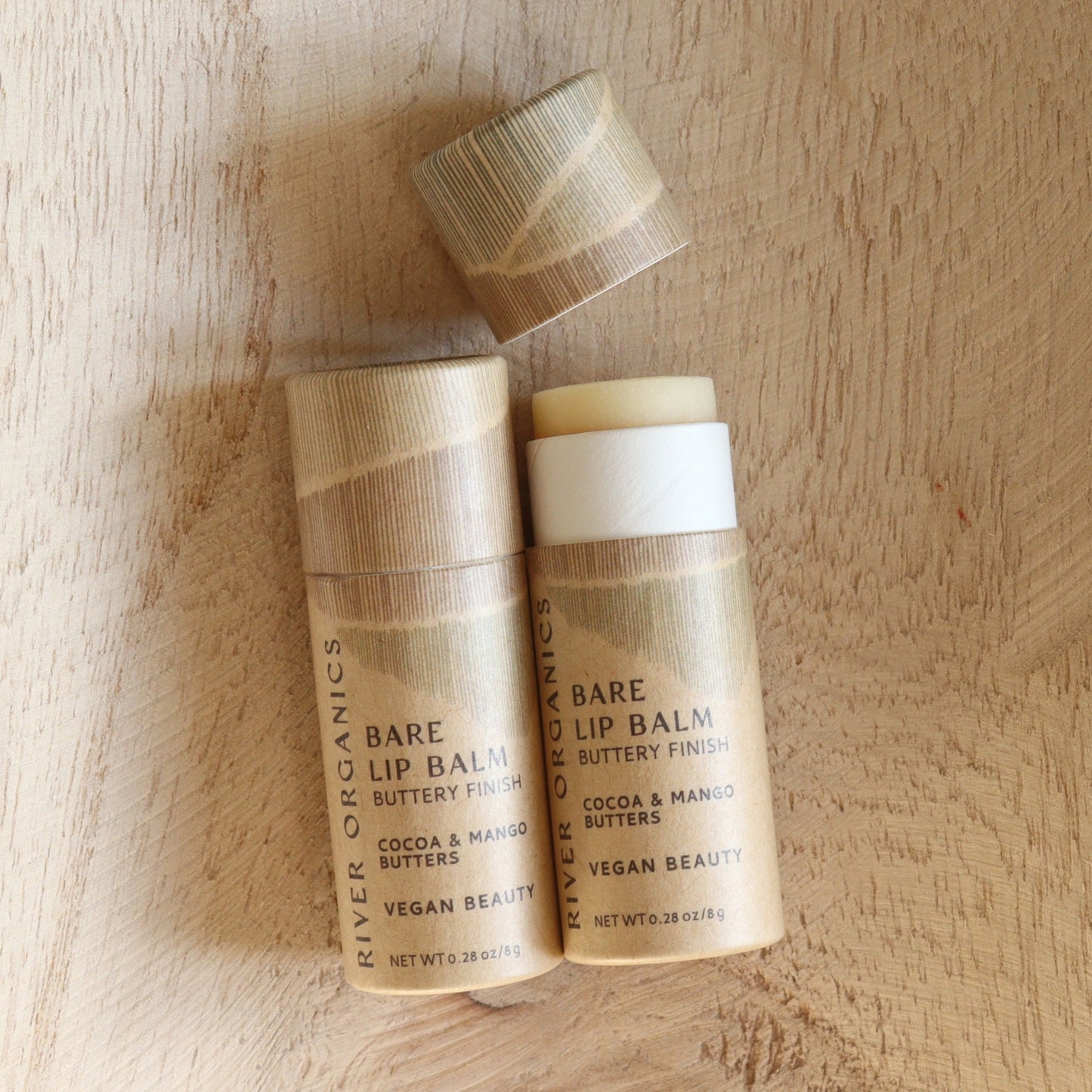 Two tubes of River Organics Bare Vegan Lip Balm rest on a wooden surface. One is closed, the other open to reveal hydrating balm with cocoa and mango butters. Both showcase eco-friendly packaging.