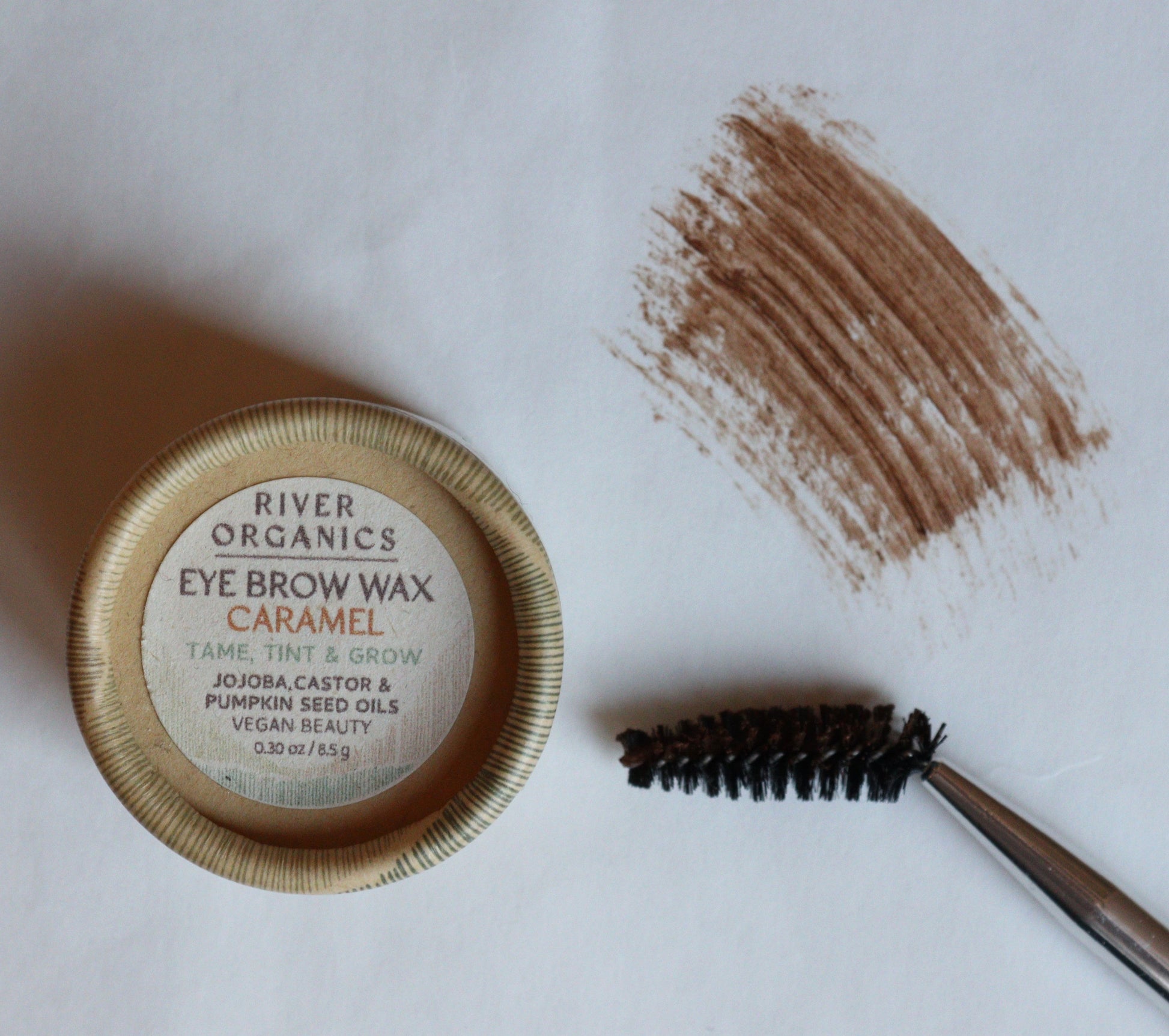A container of River Organics Brow Wax & Growth Serum sits next to a spoolie brush with brown wax on its bristles and a smear of the wax on a white surface.