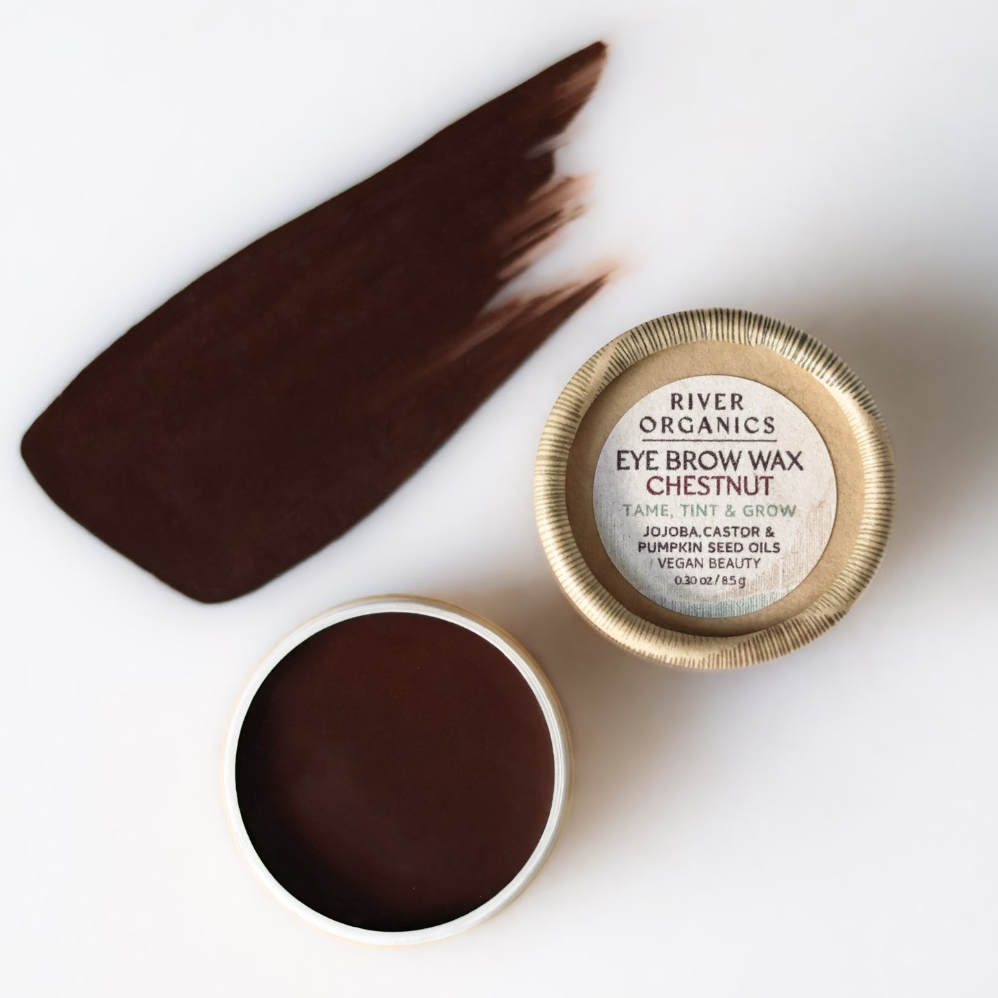 A small open tin of dark brown Imperfect Brow Wax & Growth Serum by River Organics sits next to its lid, with a swatch of the vegan brow product smeared on a white surface.