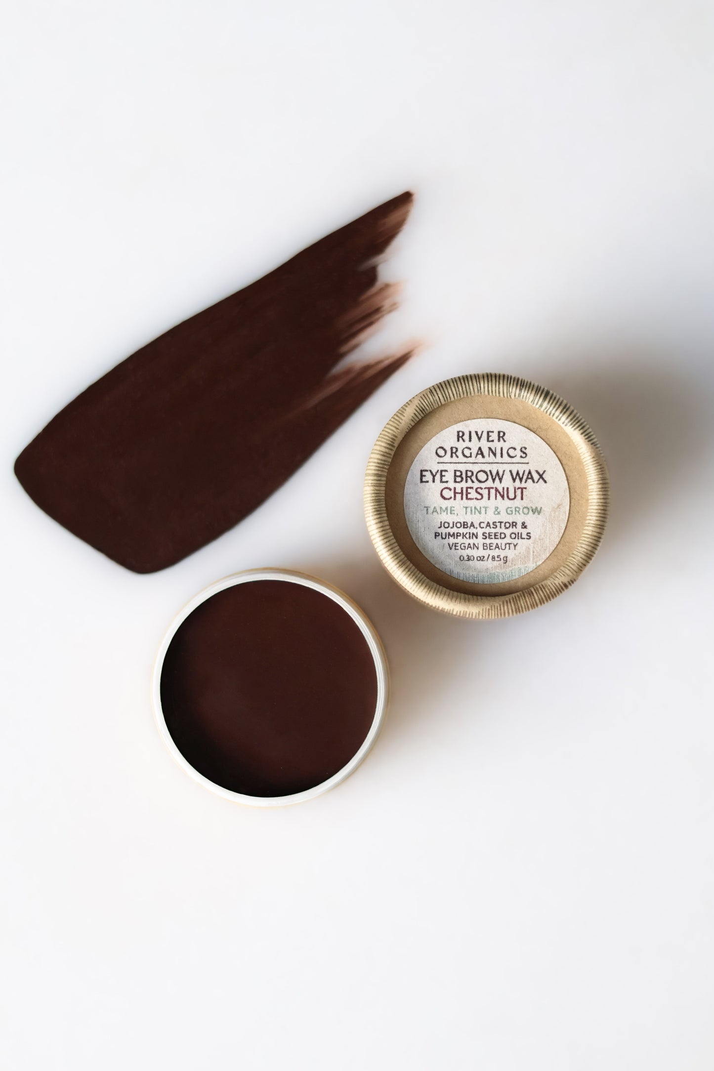 Open tin of River Organics Brow Wax & Growth Serum in Chestnut, showing a smear of dark brown brow wax on a white background with the product label visible on the lid.