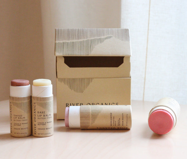 The River Organics Makeup Color Set by Tone—with blush, highlighter, and two lip products—rests on a wooden surface beside an open box. One tube lies down, revealing its pink shade against a soft curtain backdrop. Ideal for sustainable makeup gifting.