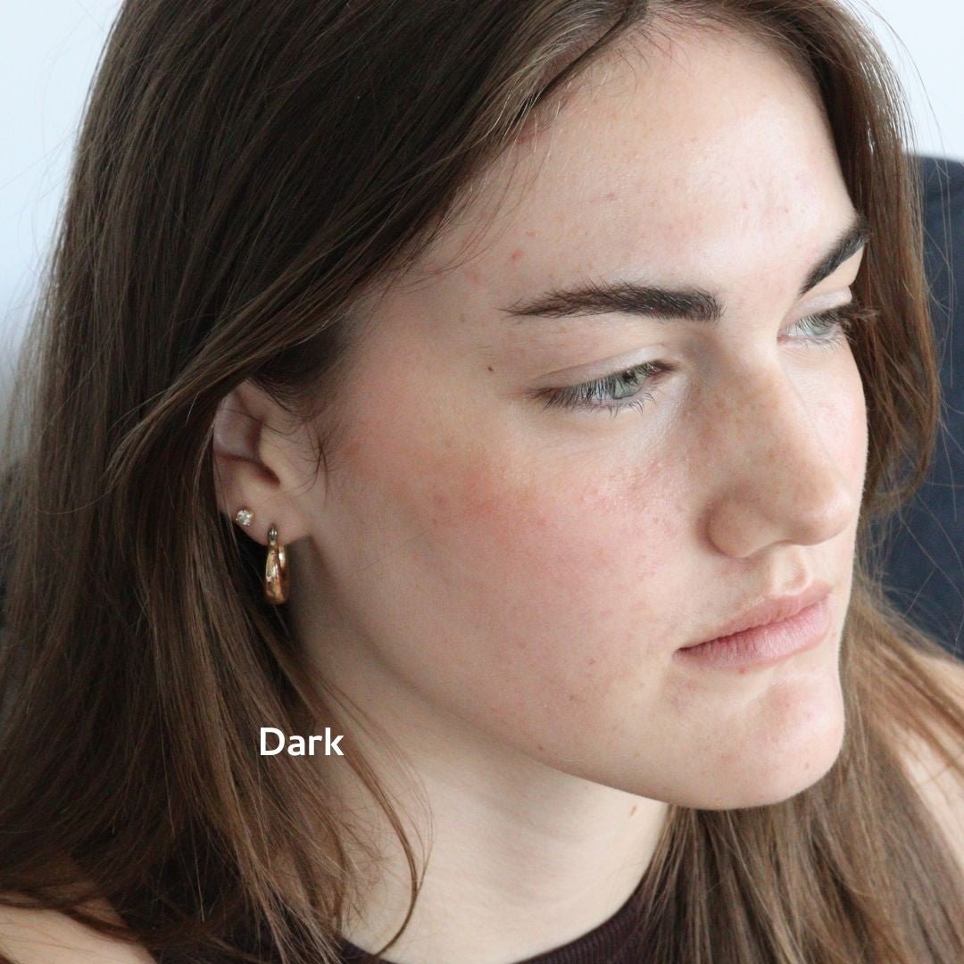 A woman with long brown hair and fair skin looks slightly away from the camera, wearing small gold hoops and River Organics Brow Wax & Growth Serum - All Colors for a polished look. The word "Dark" appears near her jawline.