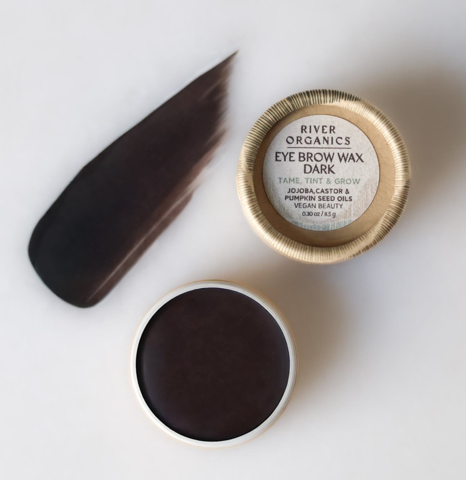 A round River Organics Brow Wax & Growth Serum in Dark is shown open with its lid beside it, revealing a smear of dark brown vegan brow product on a light surface.