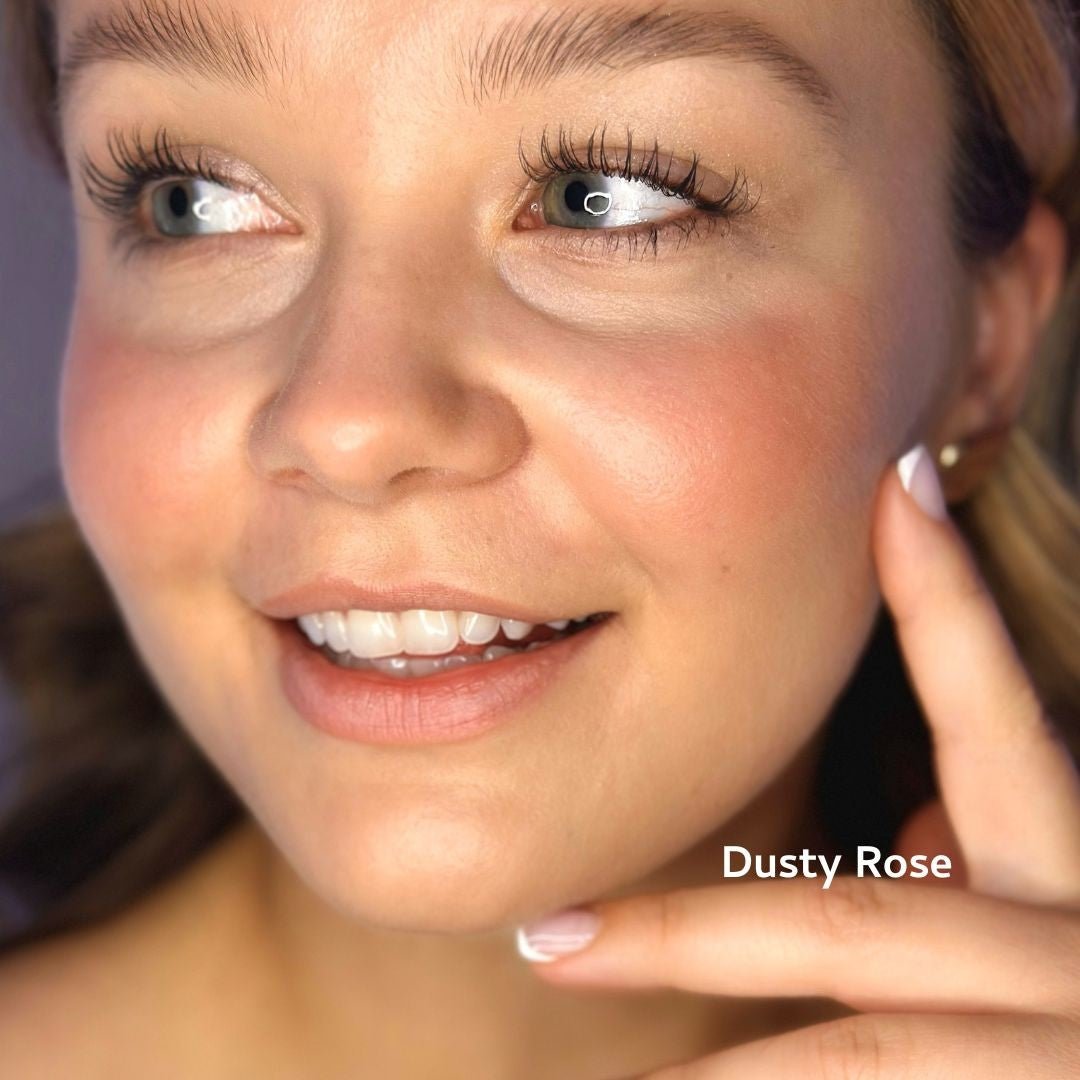 A woman with glowing skin and rosy cheeks smiles, touching her face. She wears natural makeup using River Organics Creamy Vegan Blush Sticks (Set of 2 Mix & Match) in “Dusty Rose,” highlighting a fresh, clean beauty look.