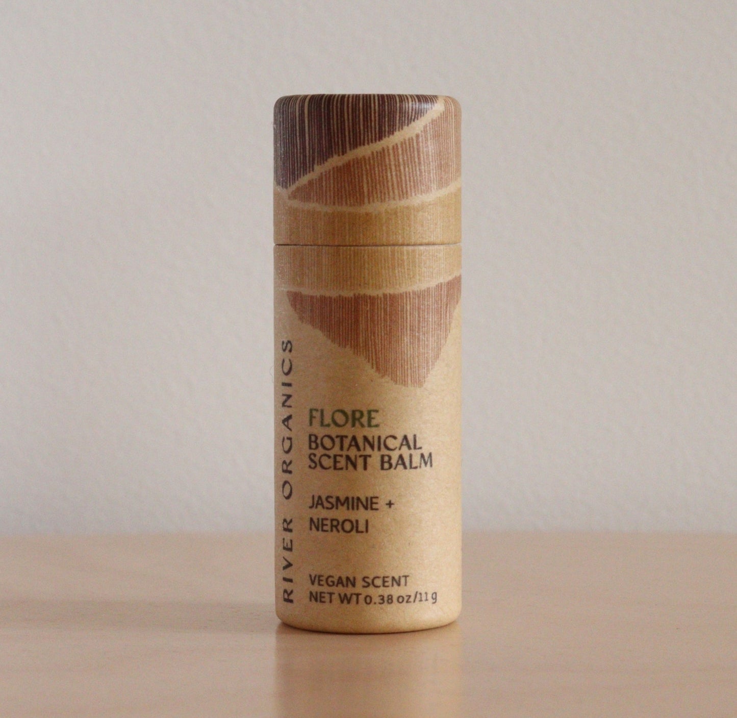 A cylindrical tube of River Organics Flore: Jasmine + Neroli - Botanical Scent Balm stands upright on a wooden surface, showcasing its natural fragrance against a plain beige background.