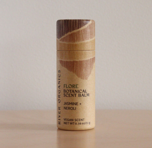 A cylindrical tube of River Organics Flore: Jasmine + Neroli - Botanical Scent Balm stands upright on a wooden surface, showcasing its natural fragrance against a plain beige background.