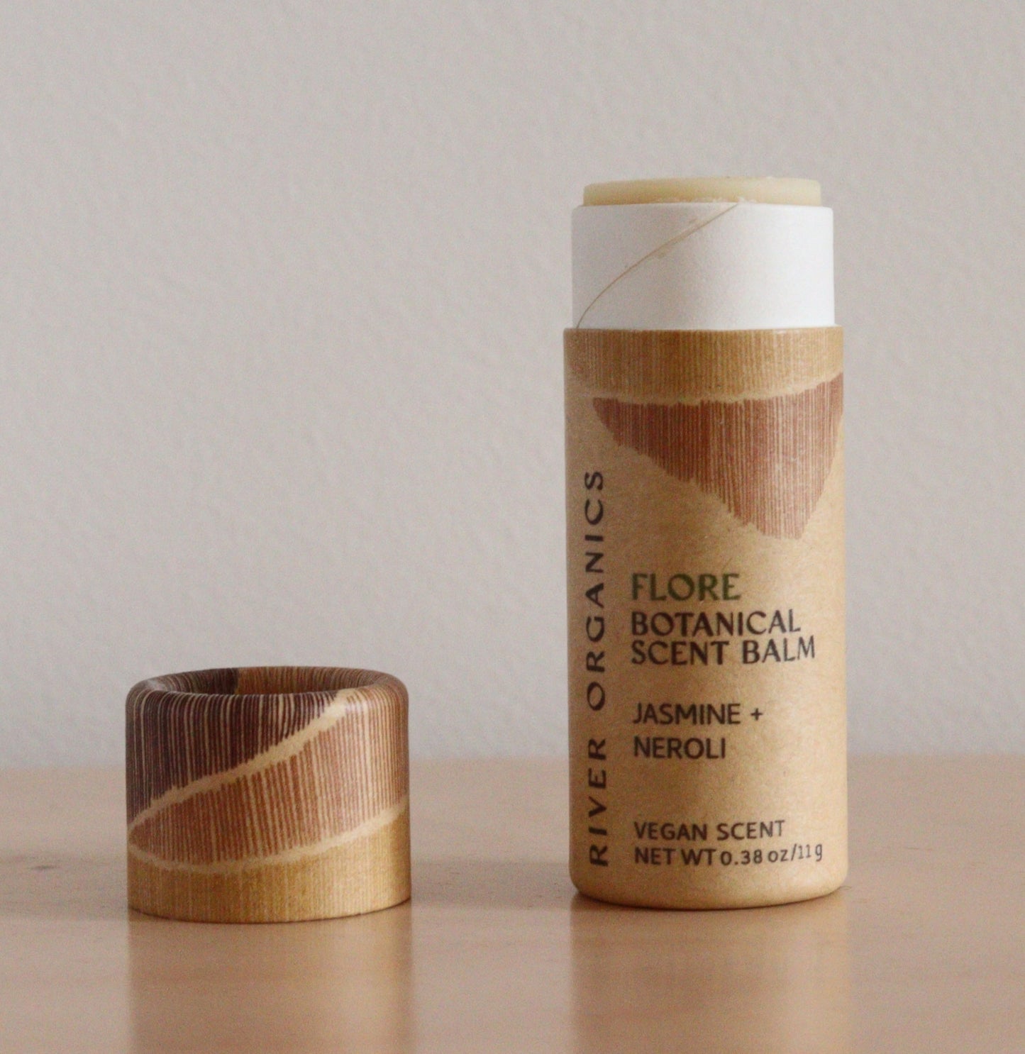 A cylindrical tube of River Organics Flore: Jasmine + Neroli - Botanical Scent Balms stands upright on wood, its cap beside it. The label features "Vegan Scent," emphasizing the product's natural fragrance.