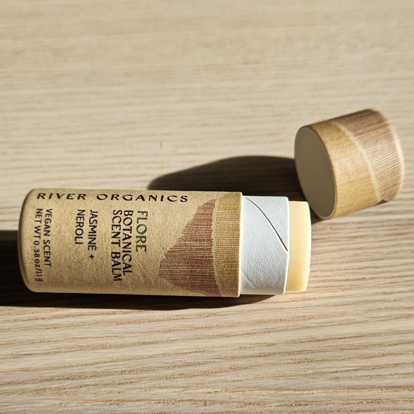 An open tube of River Organics Flore: Jasmine + Neroli Botanical Scent Balm sits on a light wood surface, its cap nearby. The brown, minimalist cardboard tube highlights this natural fragrance from River Organics.