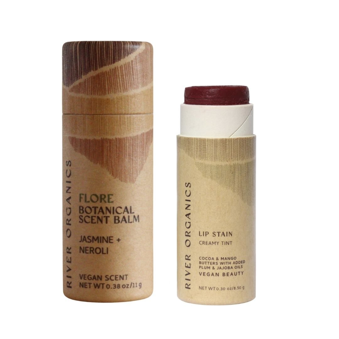 Two cylindrical cardboard tubes from River Organics’ Lip + Scent Balm Bundle are shown: a Flore Scent Balm (Jasmine + Neroli) and an open vegan lip balm with a reddish tint—both in eco-friendly packaging.