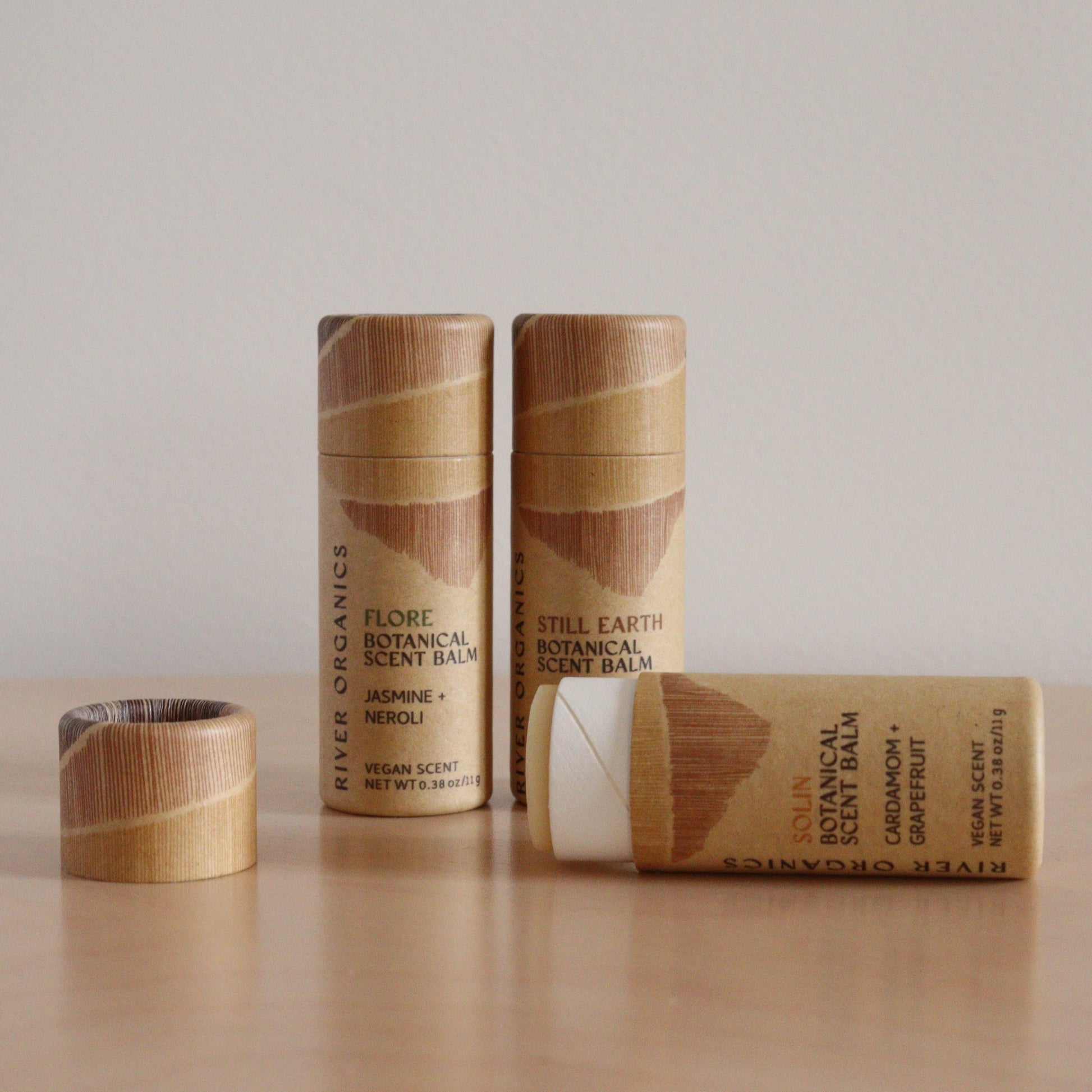 A set of River Organics Botanical Scent Balms in brown, minimalist cardboard tubes sits on wood. One tube lies open, displaying the infused balm, while the remaining tubes stand upright. Available in sets of 2, 3, or 4.