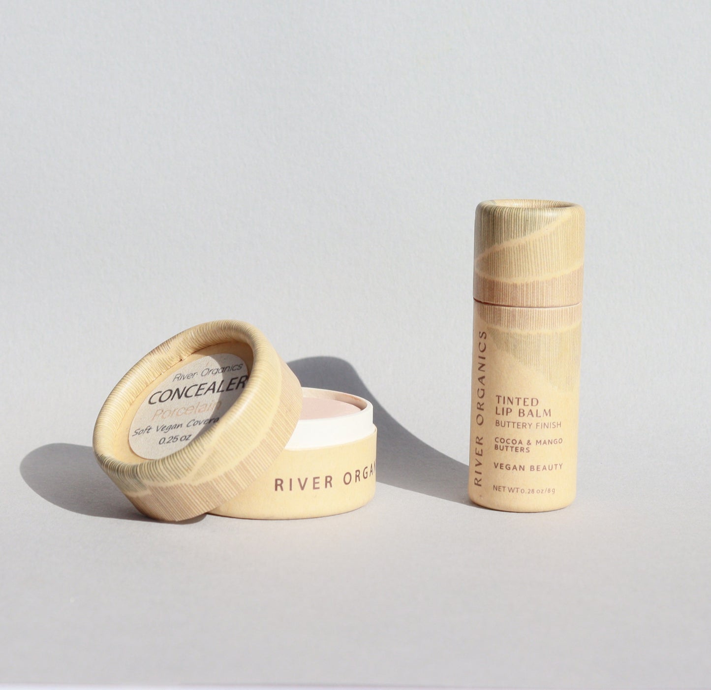 River Organics Lip + Concealer Set displayed on a light surface: an open pot of zero-waste vegan concealer with its lid leaning against it, alongside a cylindrical tube of plastic-free lip balm standing upright—both in natural-colored packaging and an ideal addition to your clean beauty routine.
