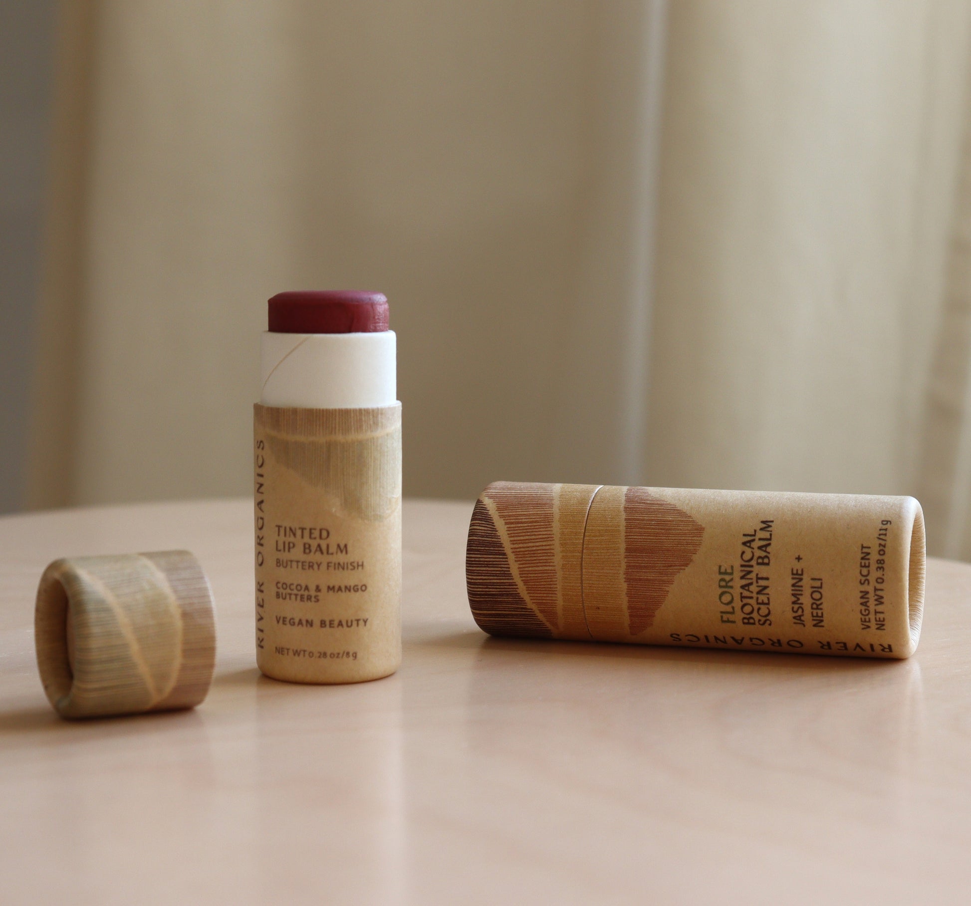 Two River Organics Lip + Scent Balm Bundle tubes rest on a table—one upright with its wooden cap nearby, another horizontal, showcasing nourishing organic oils and eco-friendly wooden packaging.