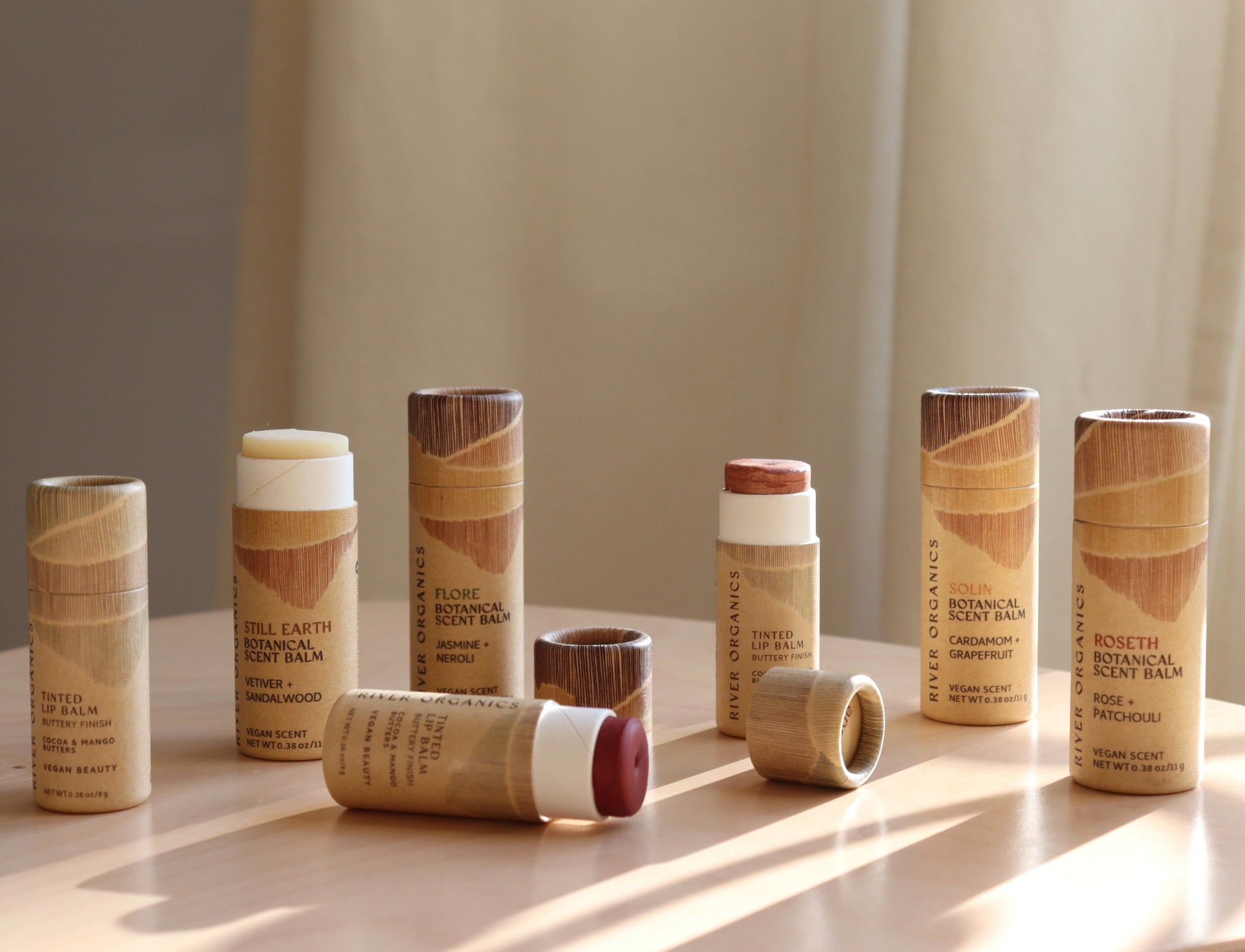 Eight cylindrical tubes of River Organics Lip + Scent Balm Bundle, some open to show the product, sit on a light wooden surface in warm sunlight. Earthy-toned, minimalist packaging evokes botanical oils and a natural wooden look.