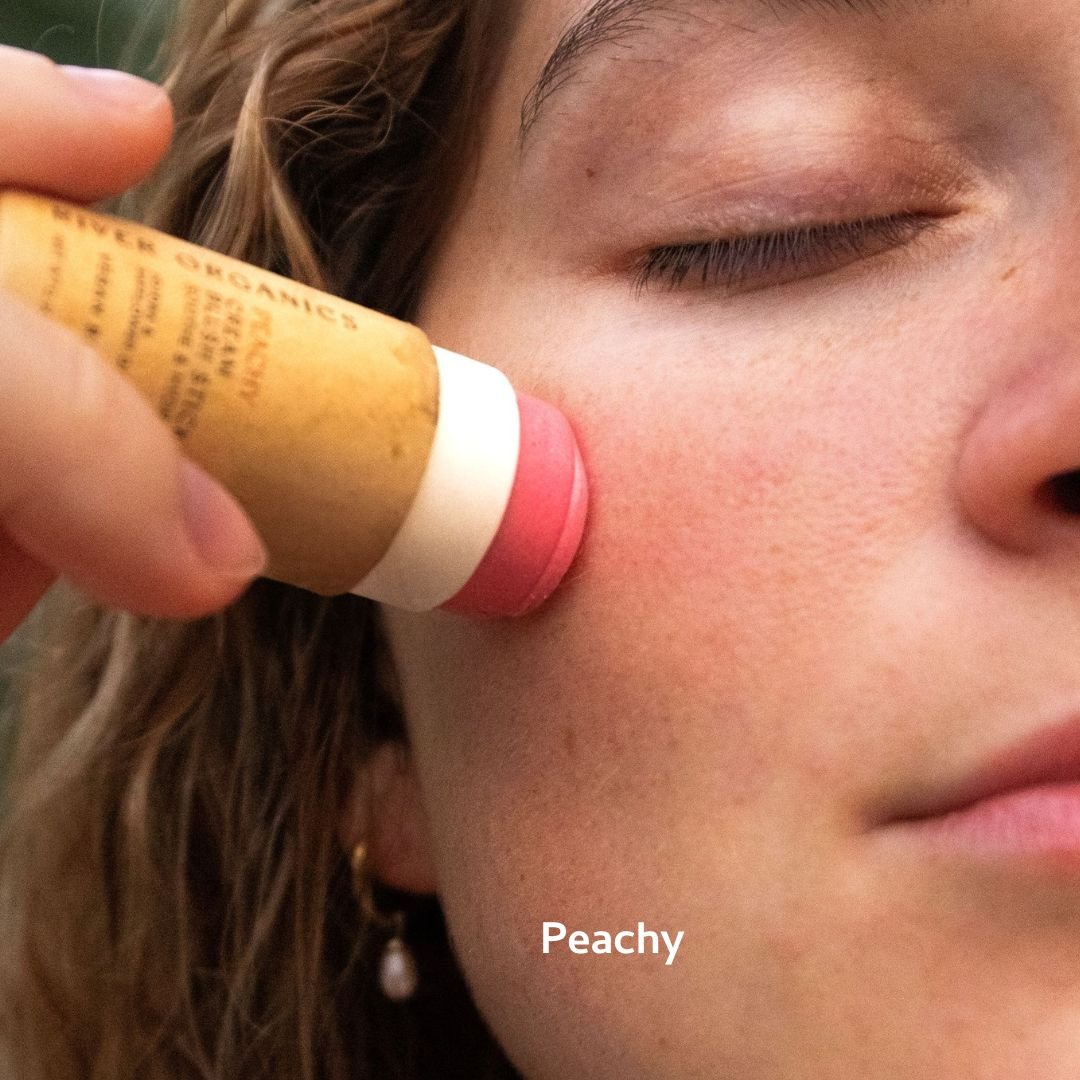 With eyes closed, a person applies River Organics' Natural Cream Blush Stick in a peach shade to their cheek. The word "Peachy" appears at the bottom of the image.
