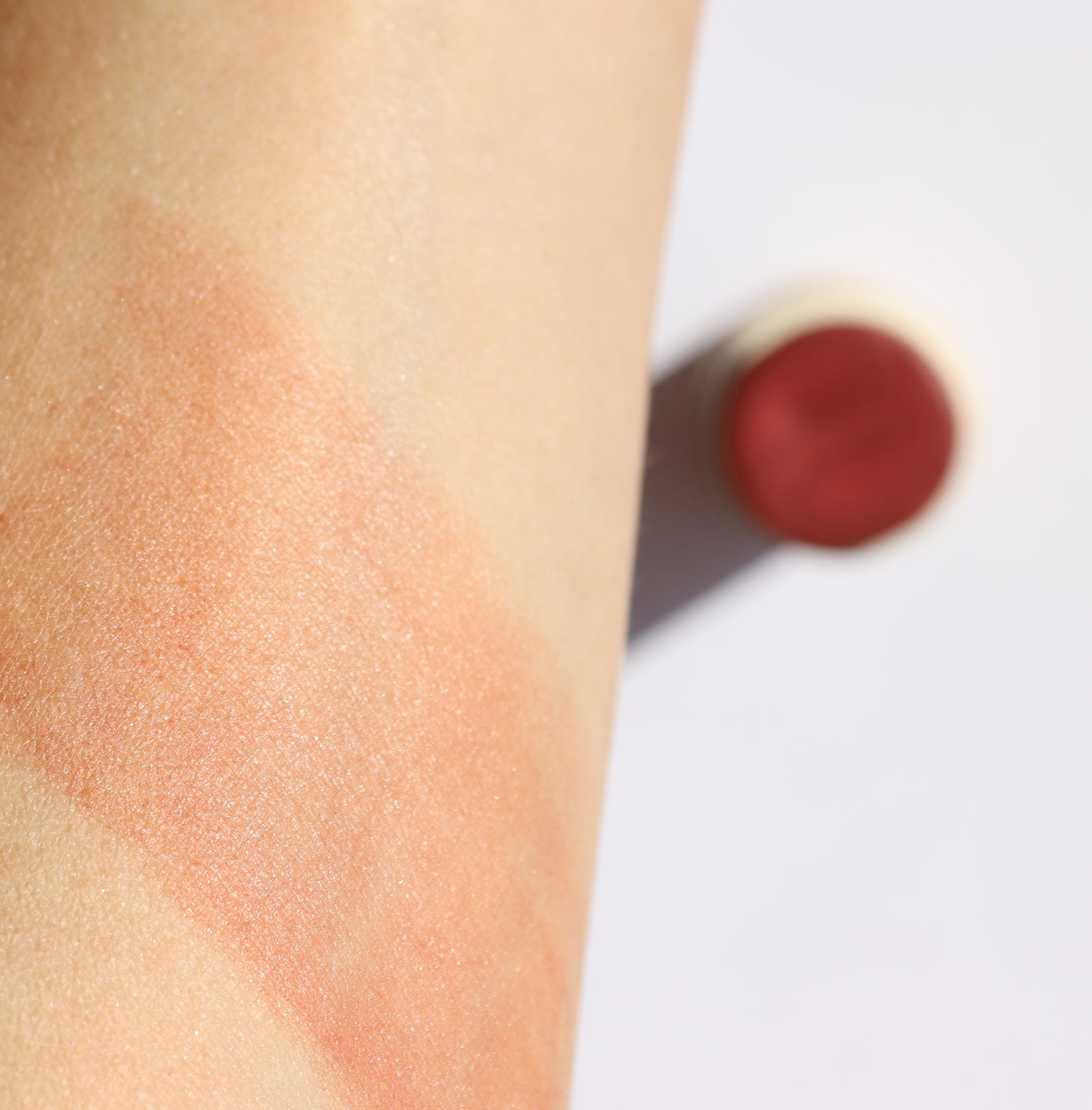 A close-up of a forearm showcases the smooth application of River Organics' Creamy Vegan Blush Stick - Pinch. The peachy, cruelty-free blush enhances the skin with nourishing ingredients. A blurred, cylindrical container is visible in the background.