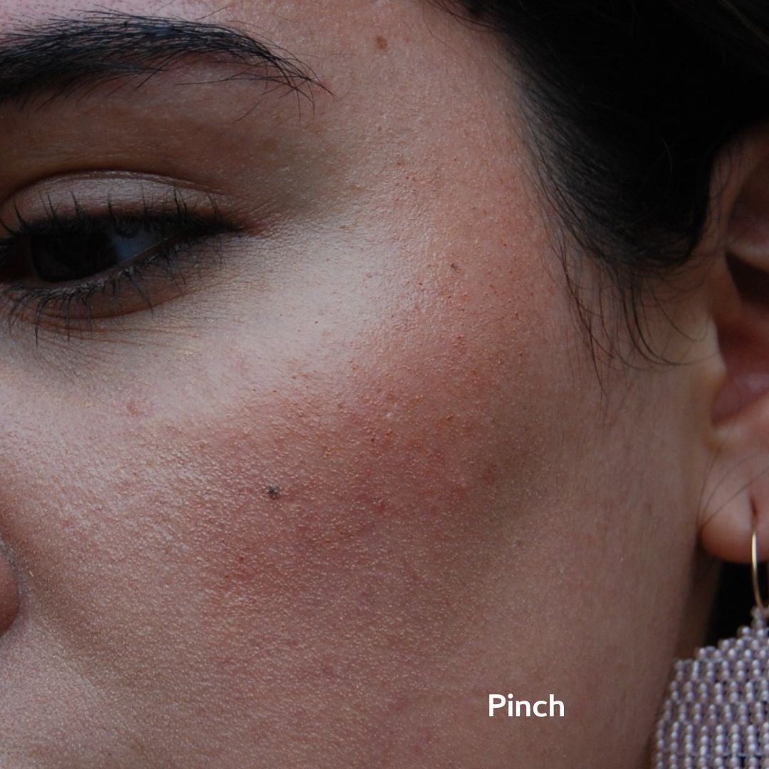 A close-up of a cheek with textured skin, visible pores, a small mole, and a sparkly earring. The person wears River Organics Natural Cream Blush Stick. "Pinch" is displayed in white text on the image.
