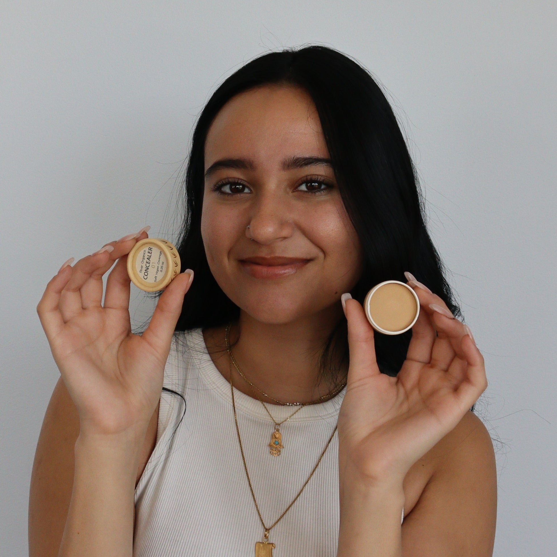 A smiling woman with long dark hair, dressed in a white sleeveless top and gold necklaces, holds up open containers of River Organics Zero-Waste Vegan Concealer (Titanium Dioxide-Free) against a plain light background.