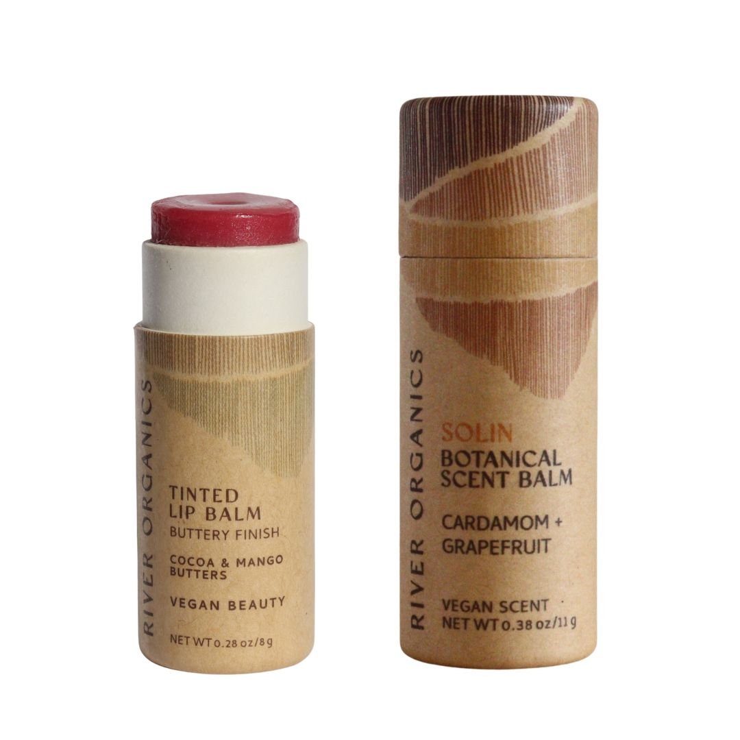 The River Organics Lip + Scent Balm Bundle features two eco-friendly, cylindrical tubes: a vegan rosy lip balm and a cardamom grapefruit scent balm, both in textured brown packaging for a botanical self-care ritual.