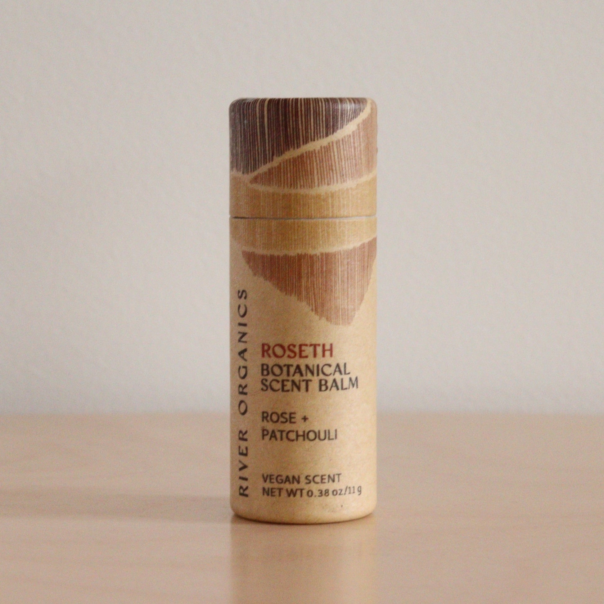 A cylindrical tube of River Organics Roseth: Rose + Patchouli - Botanical Scent Balm stands on a wooden surface against a plain light background. The beige packaging displays brown, textured graphics and text.