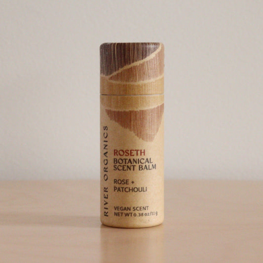 A cylindrical tube of River Organics Roseth: Rose + Patchouli - Botanical Scent Balm stands on a wooden surface against a plain light background. The beige packaging displays brown, textured graphics and text.