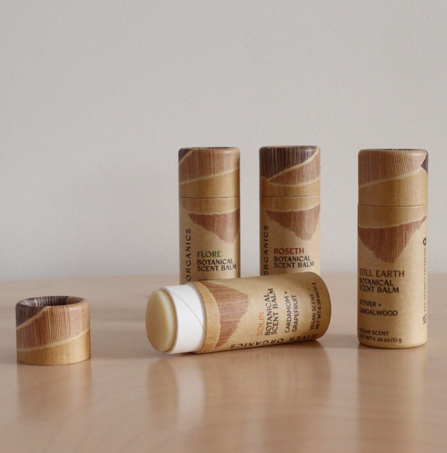 A Set of 2, 3 or 4 Botanical Scent Balms by River Organics is arranged on a light wood surface—one tube open to reveal the balm, and the others upright in their wooden-patterned packaging.