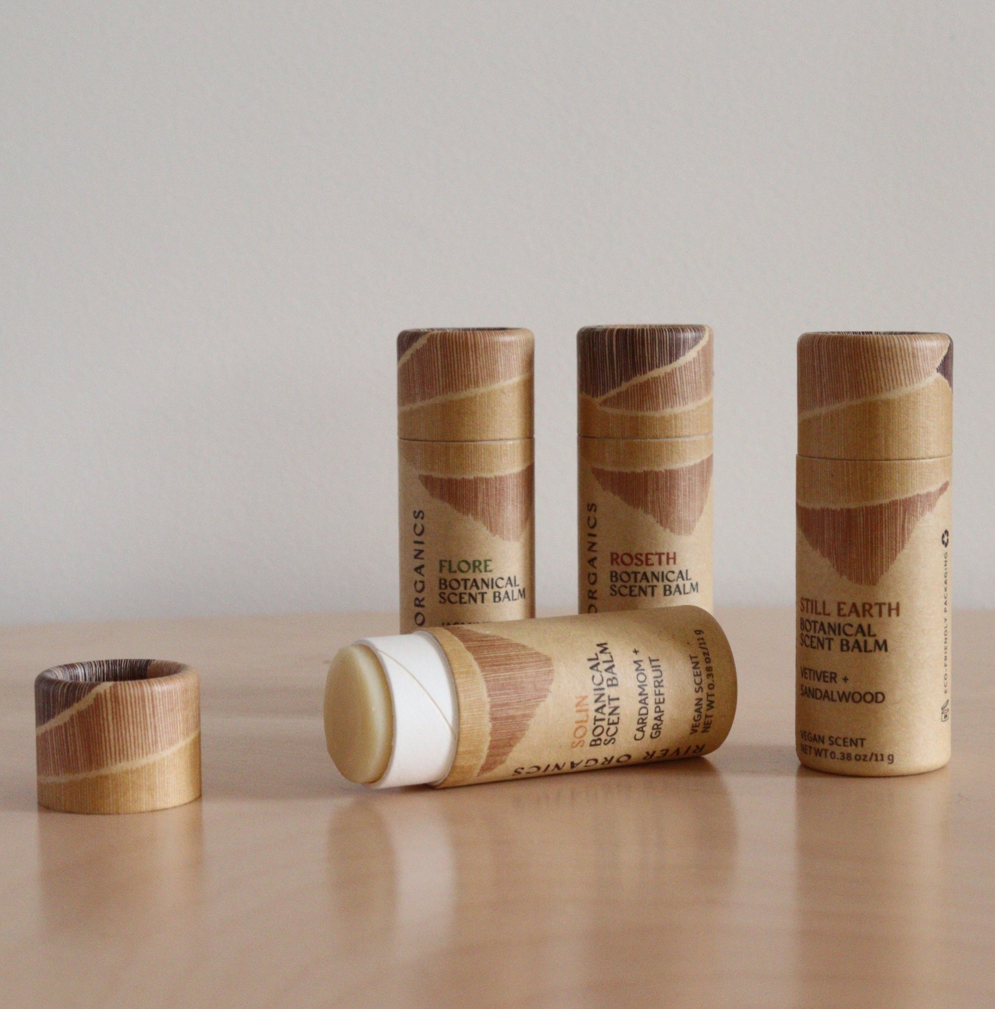 A Set of 2, 3 or 4 Botanical Scent Balms by River Organics is arranged on a light wood surface—one tube open to reveal the balm, and the others upright in their wooden-patterned packaging.