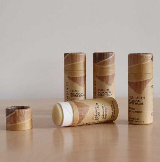 A Set of 2, 3 or 4 Botanical Scent Balms by River Organics is arranged on a light wood surface—one tube open to reveal the balm, and the others upright in their wooden-patterned packaging.