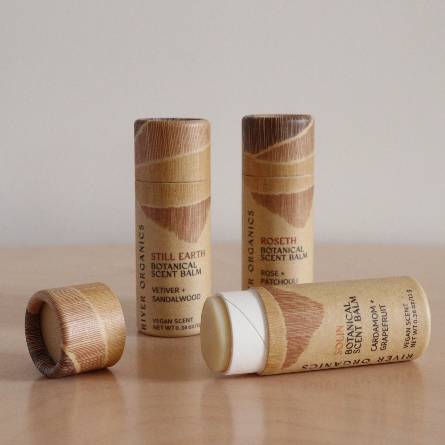 River Organics’ Botanical Scent Balms—available in sets of 2, 3, or 4—are shown with wooden caps on a light surface. One tube is open to reveal the balm, and labels feature plant-based scents including vetiver, sandalwood, rose, and patchouli.