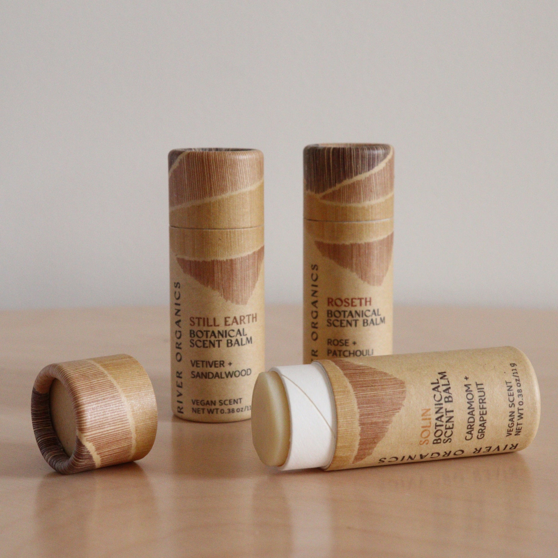 River Organics’ Botanical Scent Balms—available in sets of 2, 3, or 4—are shown with wooden caps on a light surface. One tube is open to reveal the balm, and labels feature plant-based scents including vetiver, sandalwood, rose, and patchouli.