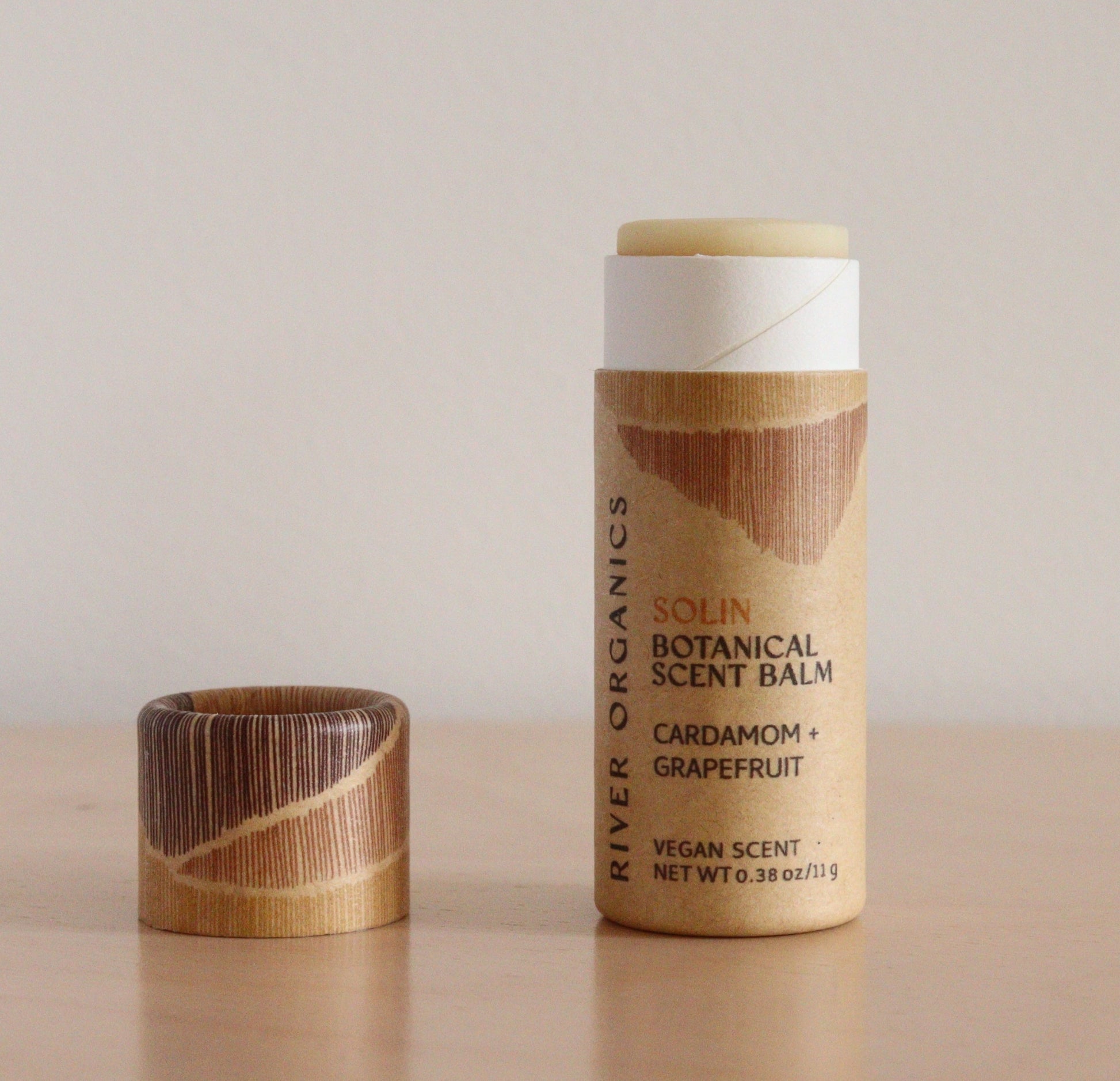 An open tube of River Organics Solin: Cardamom + Grapefruit Botanical Scent Balm, made with organic ingredients, sits on a wooden surface. The wooden cap is beside the tube, revealing the balm’s natural fragrance.