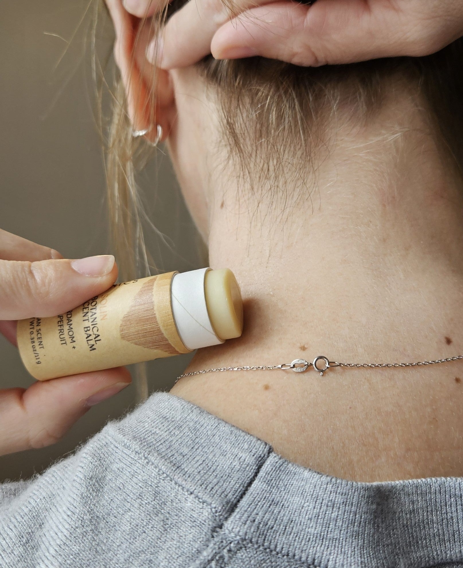 Wearing a silver chain necklace and a grey top, a person lifts their hair to apply River Organics' Solin: Cardamom + Grapefruit Botanical Scent Balm to the back of their neck.